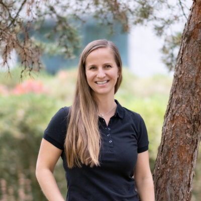Eine Frau mit langen Haaren und schwarzem Hemd lächelt, während sie im Freien in der Nähe eines Baumes steht und den Grundwert des Positiven in der Natur verkörpert.