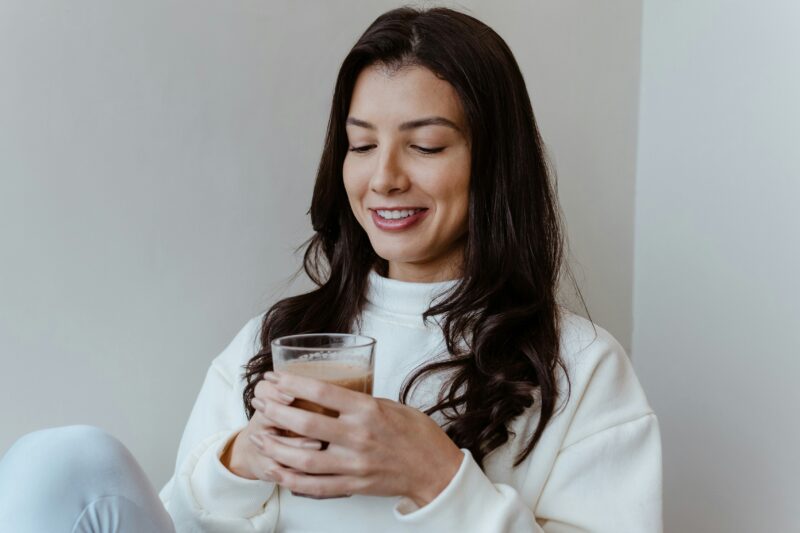 Eine Frau mit langen dunklen Haaren, die einen weißen Pullover trägt, lächelt und ein Glas mit einem braunen Getränk in der Hand hält, verkörpert wahre Gesundheit und Freude.